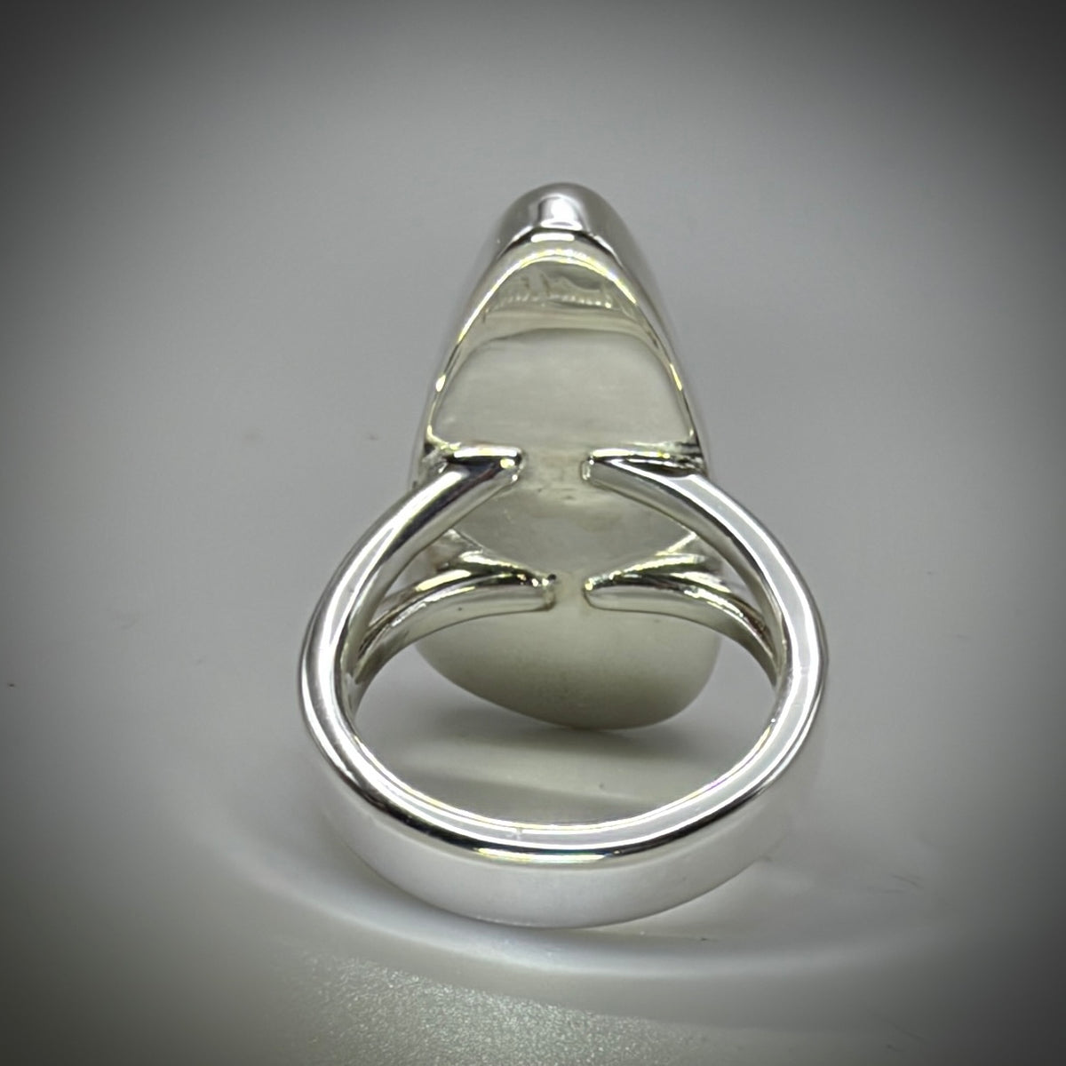 A polished silver ring with a unique triangular bezel setting holding a smooth, oval-shaped pale green stone, shown on a neutral gray background.