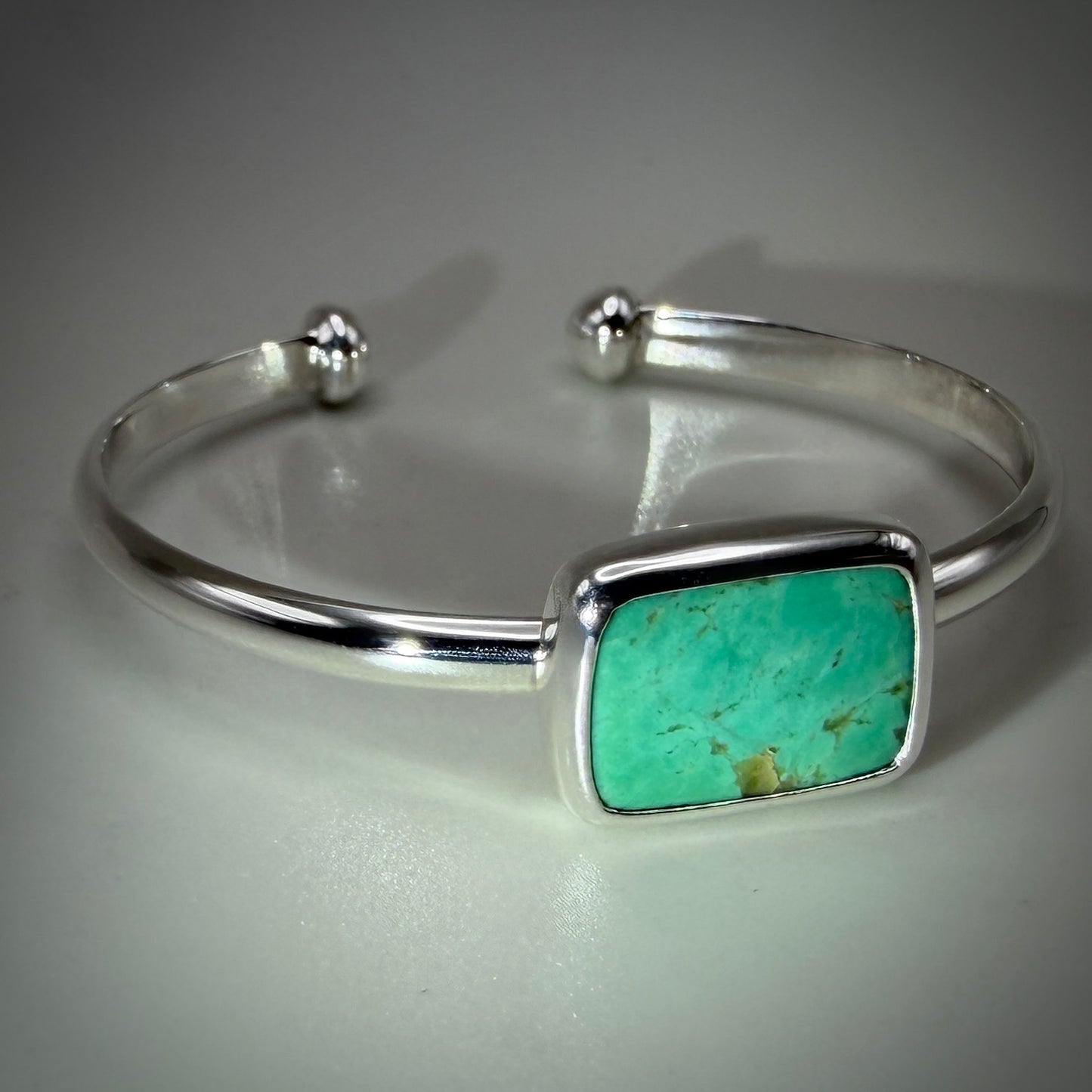 A sterling silver cuff bracelet featuring a rectangular turquoise stone inlay, displayed on a neutral gray reflective surface.