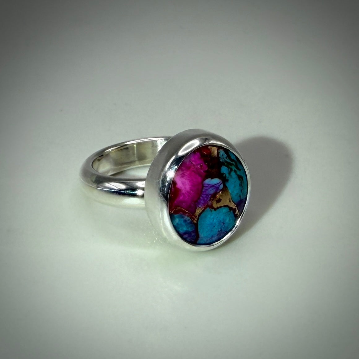 A silver band ring with a large oval bezel containing a colorful abstract pattern of turquoise blue, magenta pink, and brown, set against a neutral light gray background.