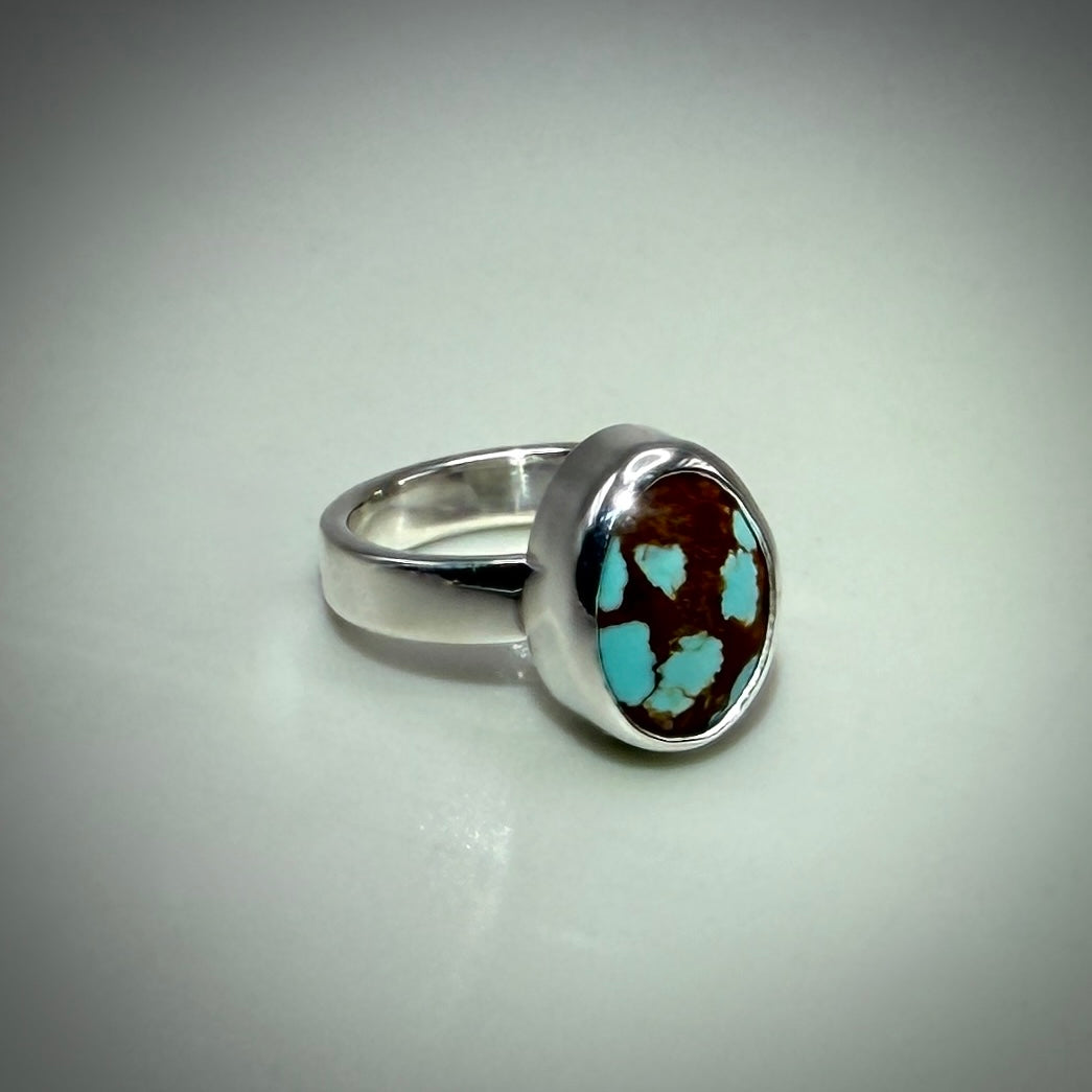 A sterling silver ring featuring a round bezel-set brown and turquoise stone, photographed on a neutral light gray background.