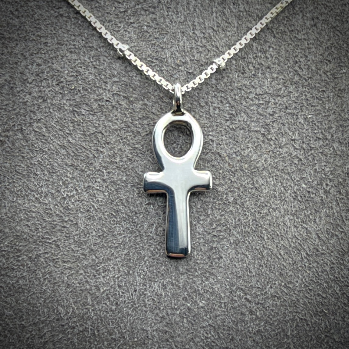 A polished silver Ankh pendant necklace on a matching silver chain, displayed against a textured gray background.