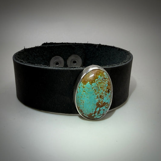 A wide black leather cuff bracelet featuring a large oval turquoise stone with brown matrix in a silver bezel setting, displayed on a neutral gray background.