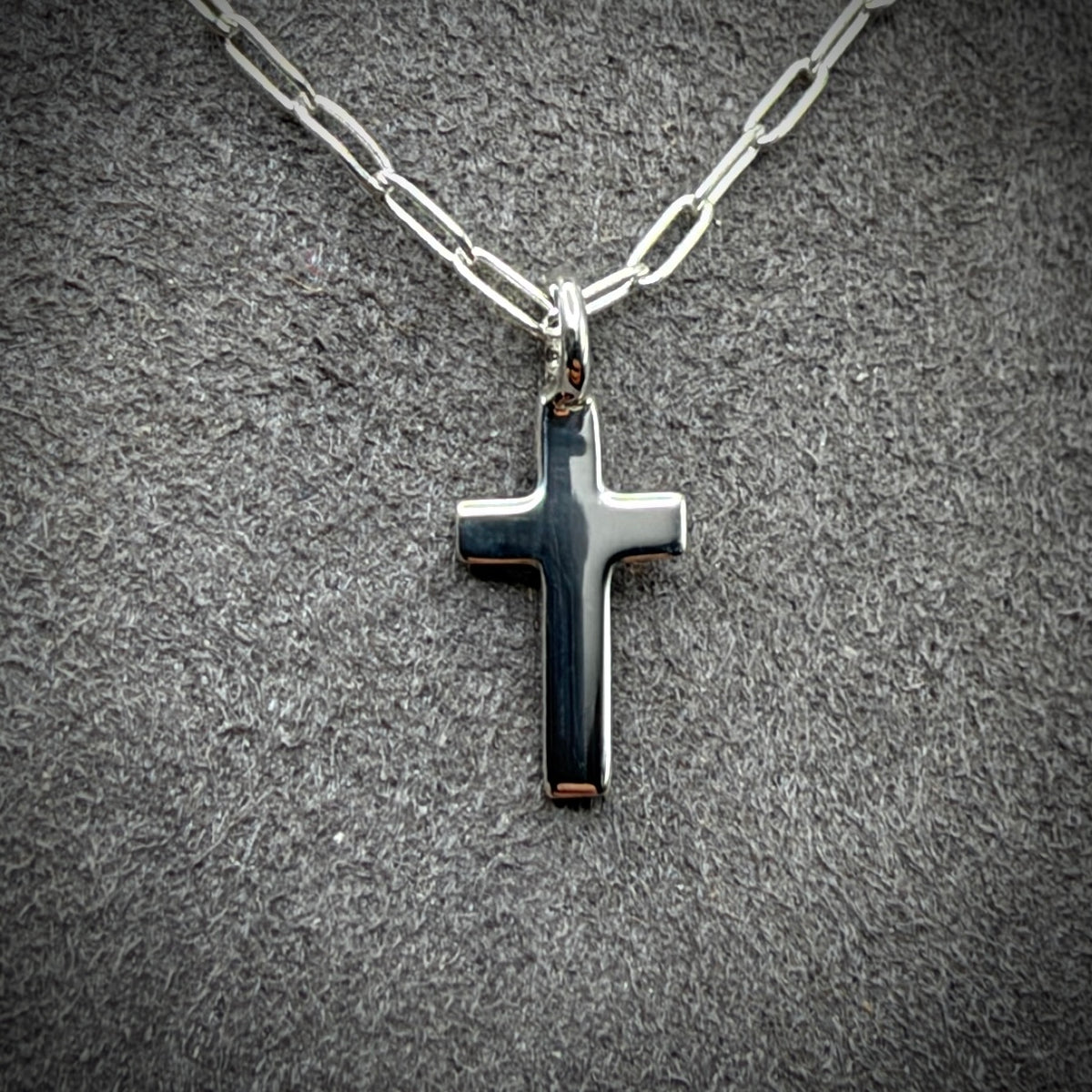 A silver cross pendant necklace with a polished finish and paperclip chain, displayed on a textured gray background.