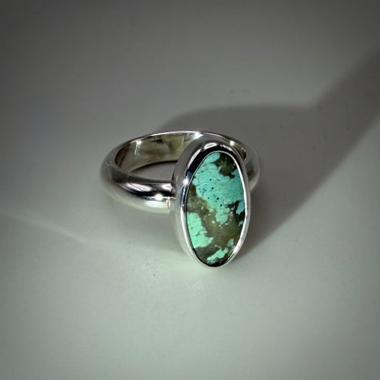 A sterling silver ring featuring an oval turquoise stone with prominent brown matrix, set against a neutral gray background.