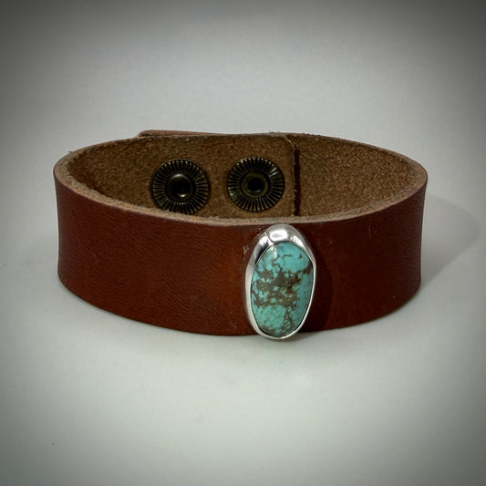 A brown leather wrap bracelet featuring a silver bezel-set oval turquoise stone on the front, with two black snap closures visible on the inside.