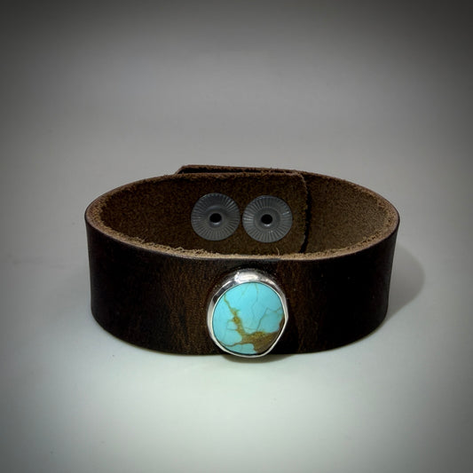 A dark brown leather cuff bracelet featuring a round turquoise stone in a silver bezel setting on the front, with two silver snap buttons on the back, shown against a neutral gray background.
