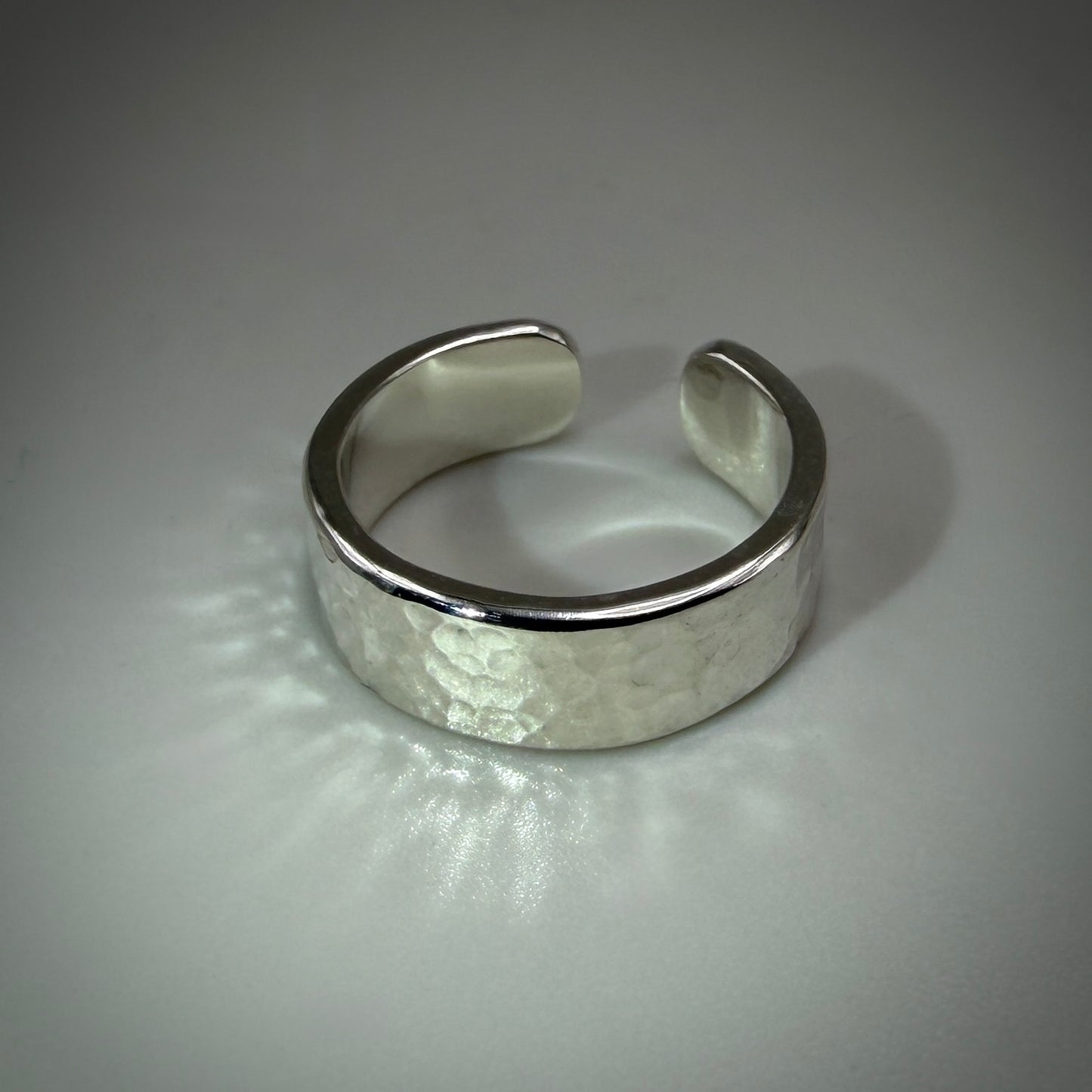 A wide silver cuff ring with a hammered texture on the front and polished beveled edges, photographed on a neutral gray background.