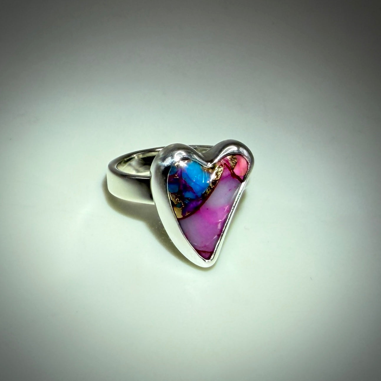 A heart-shaped silver ring featuring a vibrant inlay of pink, purple, blue, and gold marbled resin, set against a neutral gray background.
