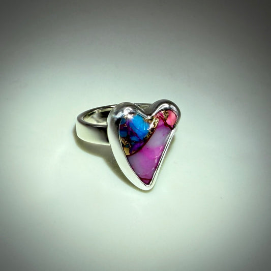 A heart-shaped silver ring featuring a vibrant inlay of pink, purple, blue, and gold marbled resin, set against a neutral gray background.