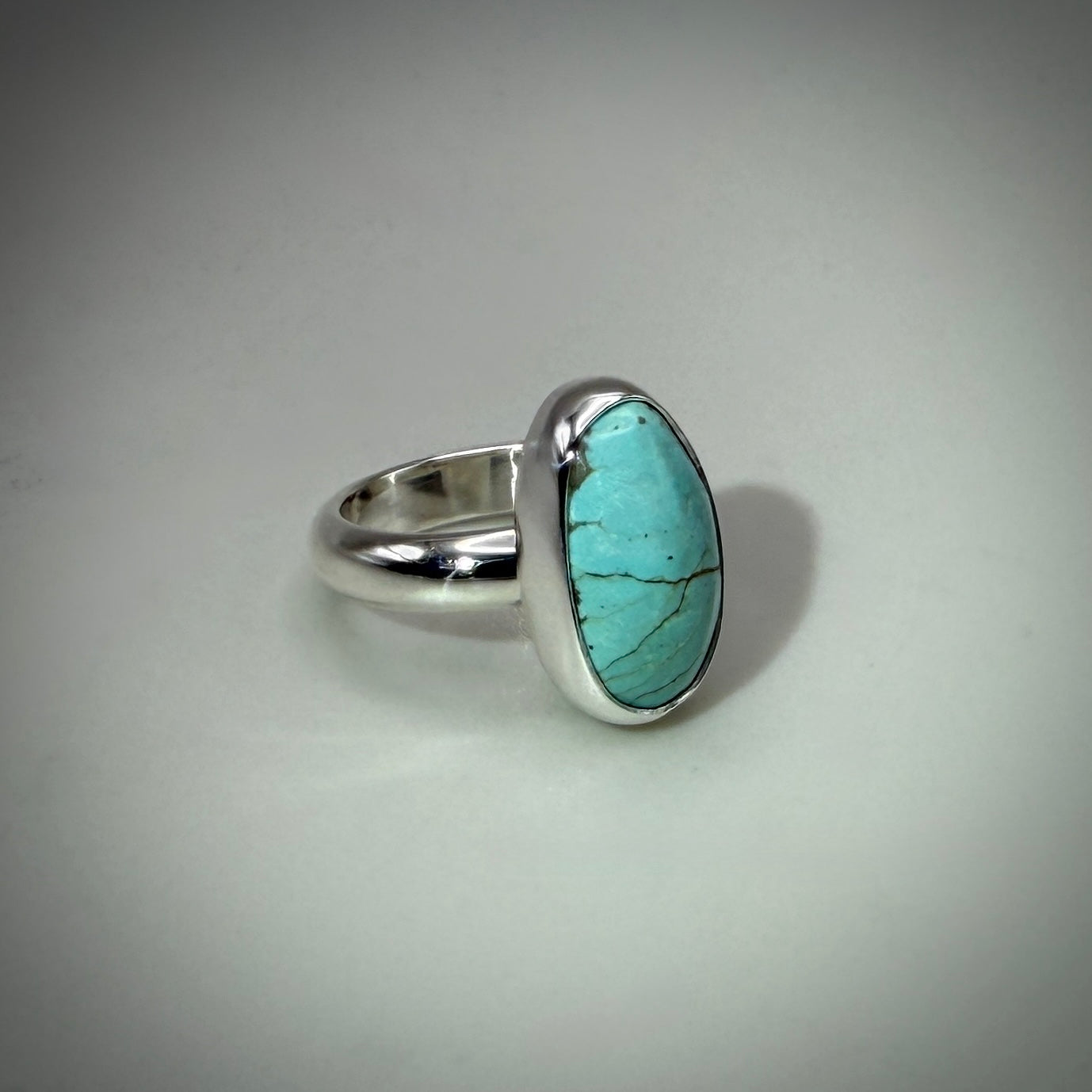 A sterling silver ring featuring an oval turquoise stone with natural matrix veining, set against a neutral light gray background.
