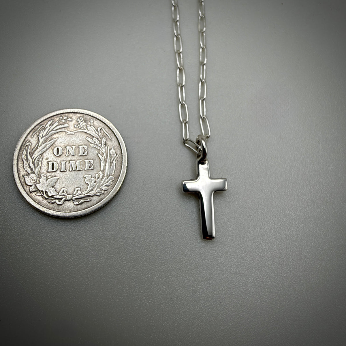 A silver cross pendant necklace on a silver chain, shown next to a silver dime coin for scale on a dark gray background.