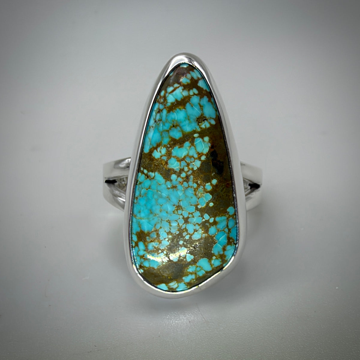 A silver ring featuring a large, teardrop-shaped turquoise stone with brown matrix and golden flecks, set against a neutral gray background.