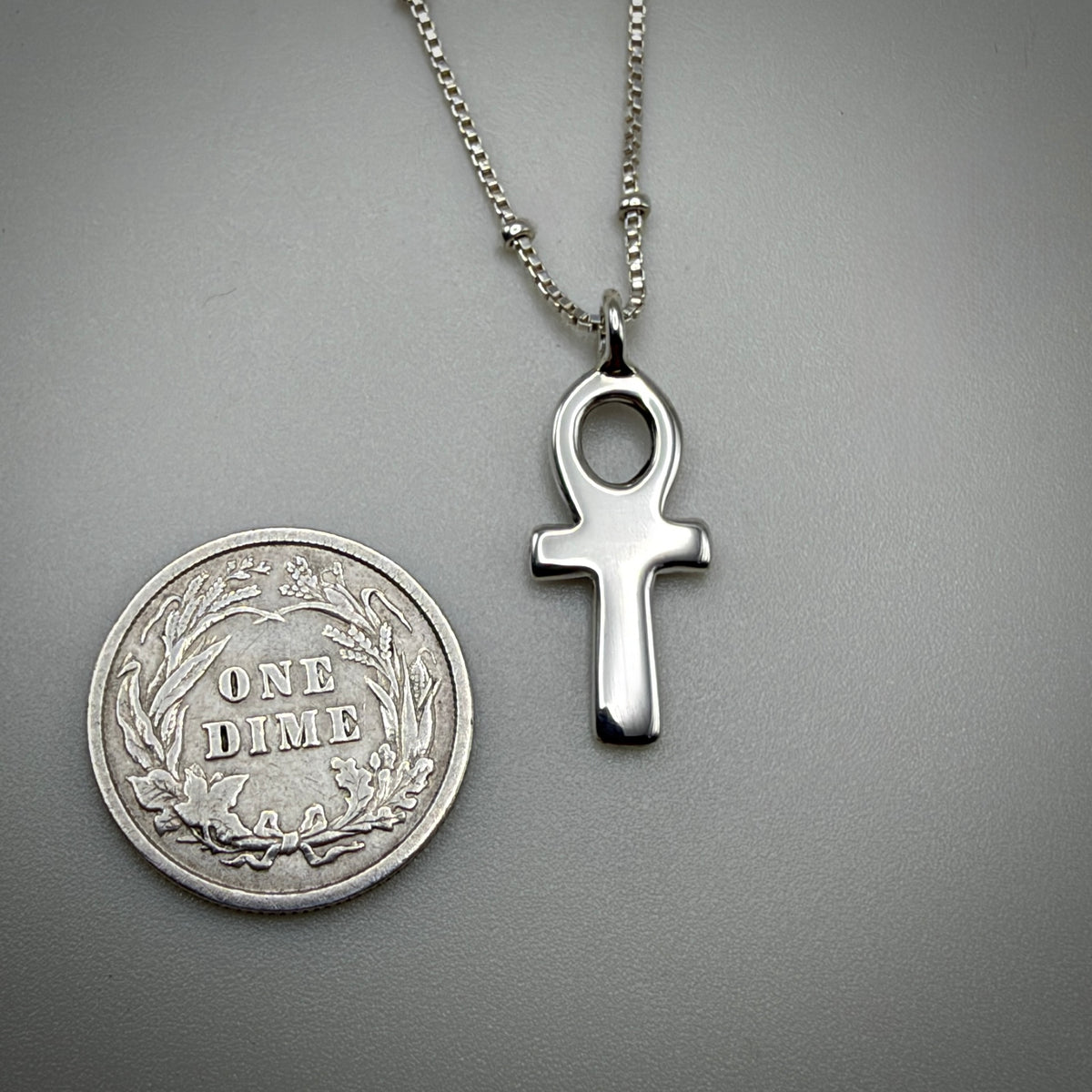 A silver ankh cross pendant necklace on a matching silver chain, displayed next to a vintage US one dime coin for scale on a neutral gray background.