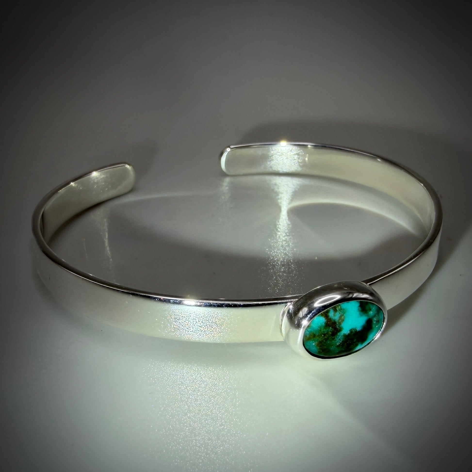 A polished silver cuff bracelet featuring a bezel-set oval turquoise stone with dark matrix, displayed on a reflective gray surface.