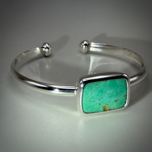 A sterling silver cuff bracelet featuring a rectangular turquoise stone inlay, displayed on a neutral gray reflective surface.