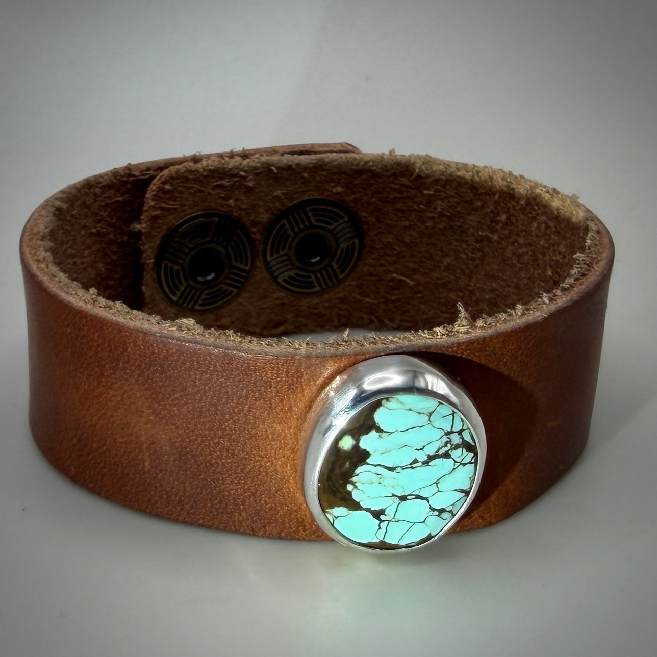 A brown leather cuff bracelet featuring a large oval silver bezel set with a turquoise-colored stone containing black veining, photographed on a neutral gray background.