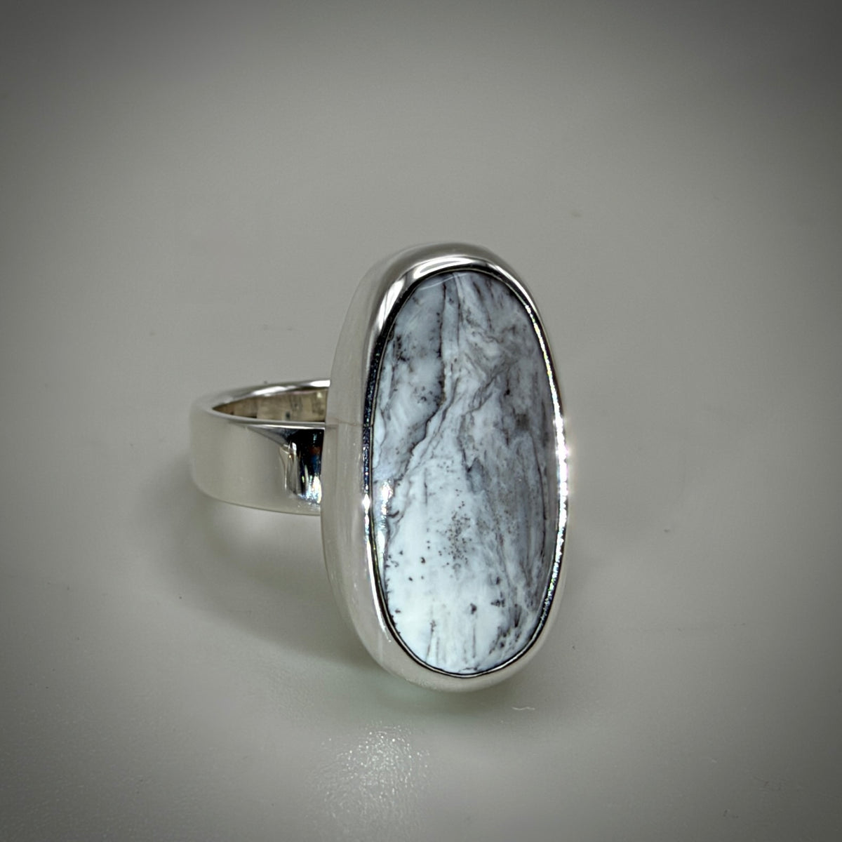 A silver ring featuring a large, oval-shaped cabochon stone with a white and gray marbled pattern, set in a polished silver bezel on a neutral gray background.