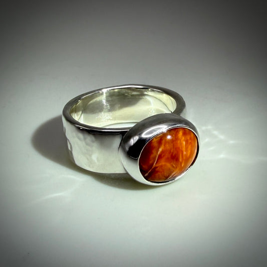 A silver-toned ring with a wide band and a round bezel-set orange-brown stone, photographed on a light gray background.