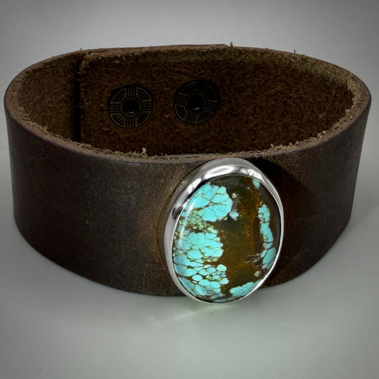 A wide brown leather cuff bracelet featuring a large oval turquoise stone in a polished silver bezel, shown on a neutral gray background.