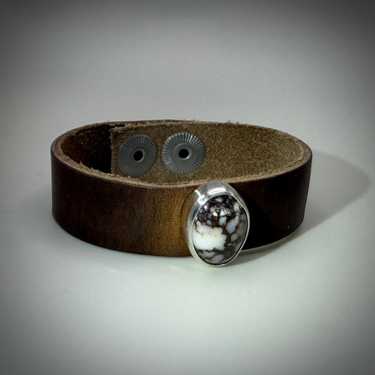 A dark brown leather cuff bracelet with a silver-toned bezel holding a white and black marbled stone, shown on a neutral gray background.