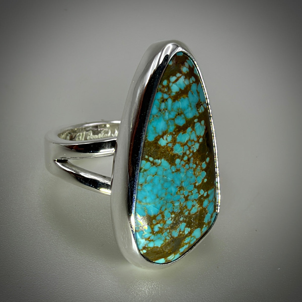 A sterling silver ring featuring a large, irregularly shaped turquoise stone with prominent brown matrix veining, set against a neutral gray background.