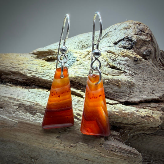 A pair of triangular dangle earrings with orange and red banded agate stone pendants on silver fishhook wires, displayed on a piece of weathered driftwood.