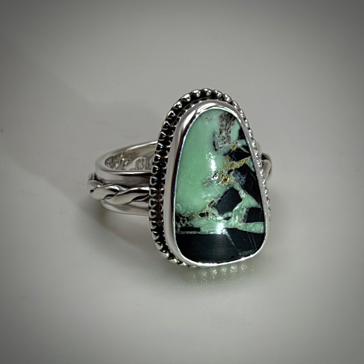 A silver ring featuring a large, irregular-shaped green stone with black and gold matrix patterns, set in a textured silver bezel with a twisted silver band.