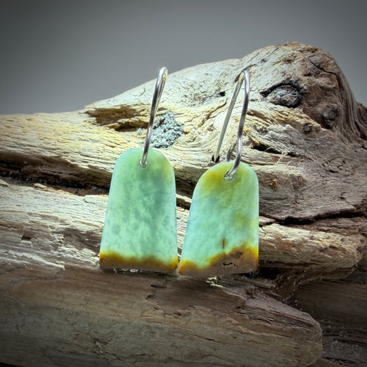 A pair of handmade earrings featuring irregular light green and yellow-green stone pendants with natural brown edges, suspended from silver fishhook wires, displayed on a piece of weathered driftwood.
