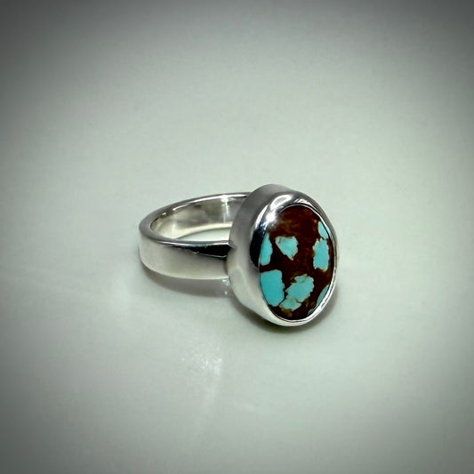 A sterling silver ring featuring a round bezel-set brown and turquoise stone, photographed on a neutral light gray background.