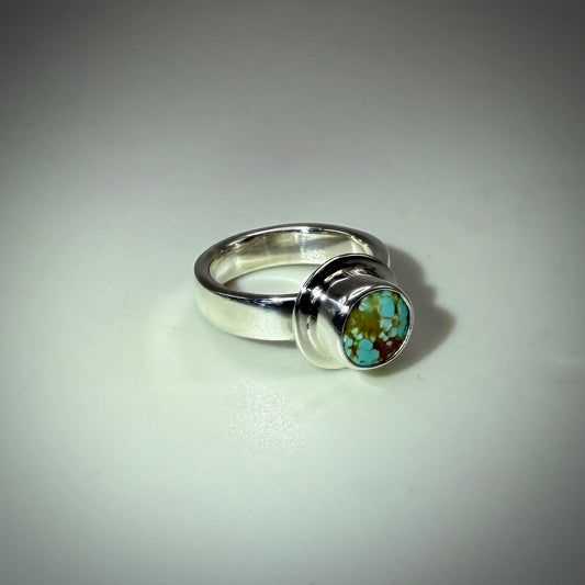 A sterling silver ring featuring a round bezel-set turquoise stone with visible brown matrix, photographed on a neutral light gray background.