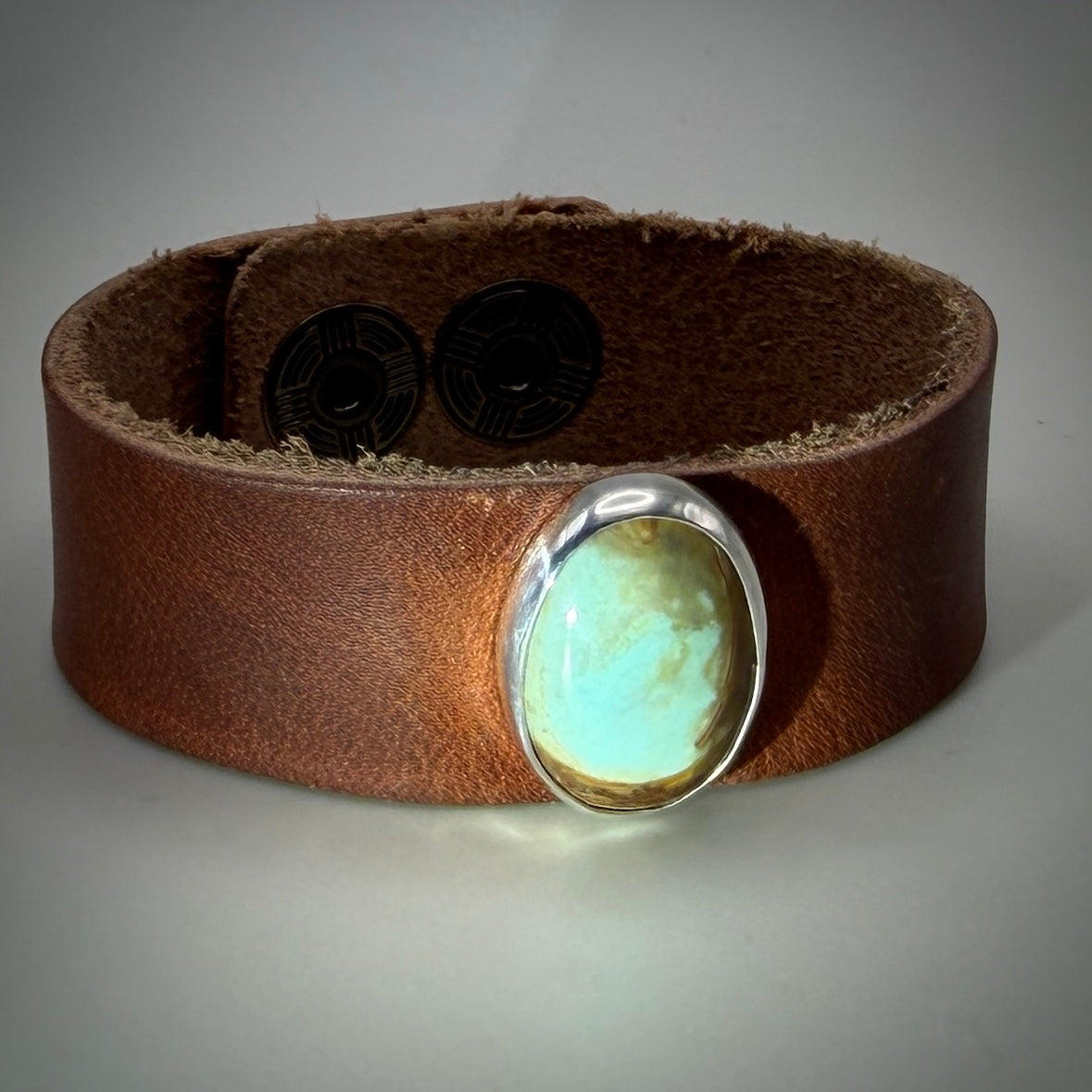 A brown leather cuff bracelet featuring a large oval turquoise stone set in a polished silver bezel, displayed on a neutral gray background.