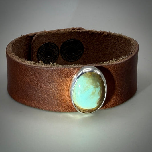 A brown leather cuff bracelet featuring a large oval turquoise stone set in a polished silver bezel, displayed on a neutral gray background.